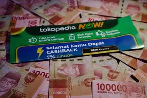 Jakarta, Indonesia in December 2022. A Tokopedia Now flyer above several hundred thousand rupiah notes scattered around. photo