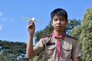 el boy scout asiático sostiene un modelo de avión blanco contra un fondo nublado y azul, enfoque suave y selectivo. foto