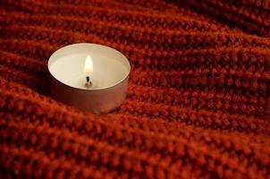Candle stands on brown knitted cloth. Ginger color knitted cloth. Warm colors photo. Autumn and winter. Cold season concept. Cozy and comfortable concept. One burning candle. Knitted material texture. photo