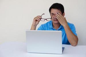 Asian man, feels hurt his eyes, holds eyeglasses during working on laptop. concept , Eyesight problem. Optometry. Glasses with convex or concave lenses. Myopia.Eye disease photo