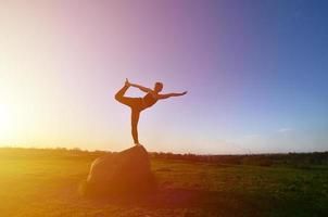 la silueta de una joven rubia con traje deportivo practica yoga en una pintoresca colina verde por la noche al atardecer. el concepto de ejercicio y estilos de vida saludables foto