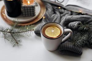 Winter home cozy concept. Mug with lemon tea, open book, warm sweater, candles and fir tree. Wellbeing, relaxing concept photo