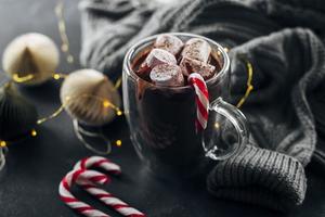 vaso con chocolate caliente con malvavisco y bastón de caramelo sobre fondo azul oscuro. foto