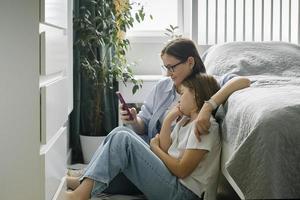 madre e hija usando un teléfono inteligente mientras están sentados en berdoom. mujer y niña preadolescente juntas foto