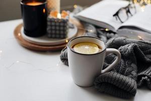 Winter home cozy concept.Cup of tea with lemon, open book, warm sweater, candles and fir tree. Wellbeing, relaxing concept photo