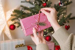 Female hand decorating gift box near christmas tree. Festive colorful background