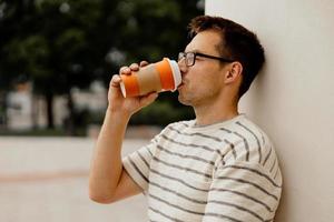 Young adult man sitting outdoors, drinking coffee, enjoying good weather and city view. Man has a break from work, spending time outside and relaxing. Time with yourself, dreaming, mental health. photo