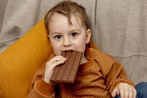 Little adorable boy sitting on the couch at home and eating chocolate bar. Child and sweets, sugar confectionery. Kid enjoy a delicious dessert. Preschool child with casual clothing. Positive emotion. photo