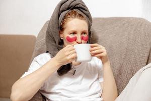 Young woman with casual clothes, towel on head and eye patches drinking hot tea and resting. Relax after taking shower. Cosmetic for women, skin care. Self care, time for yourself. Relaxing at home. photo