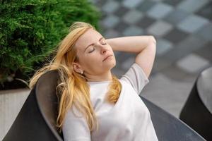 Young beautiful blond woman sitting outdoors, on the wooden terrace in the city and resting. Girl has break, spending time outside and relaxing. Time with yourself, dreaming, relaxation, mental health photo