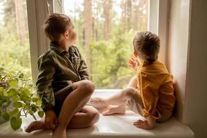 niños sentados en el alféizar de la ventana y esperando que alguien venga. dos hermanos, amigos. lindos niños en edad preescolar solos en casa. los niños están esperando a su madre o padre. soledad. padres ocupados. foto