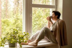 Middle age beautiful woman sitting on windowsill, drinking tea, dreaming. 50-year-old woman relaxing with cup of tea. Relax at home, looking away, thinking, stress free, peaceful mood, wellbeing alone photo