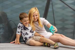 Happy blond woman and little boy sitting on terrace and making selfie on smartphone. Mother and son enjoy time together. Positive young mom spending time with her cute child. Having fun. Family. photo