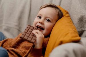 Little adorable boy sitting on the couch at home and eating chocolate bar. Child and sweets, sugar confectionery. Kid enjoy a delicious dessert. Preschool child with casual clothing. Positive emotion. photo