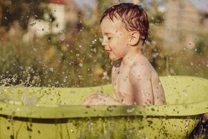 Cute little boy bathing in tub outdoors in garden. Happy child is splashing, playing with water and having fun. Summer season and recreation. Staying cool in the summer heat. Water fun in backyard. photo