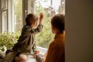 niños sentados en el alféizar de la ventana y esperando que alguien venga. dos hermanos, amigos. lindos niños en edad preescolar solos en casa. los niños están esperando a su madre o padre. soledad. padres ocupados. foto