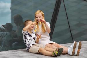 mujer rubia feliz y niño sentado en la terraza y comiendo dulces. madre e hijo disfrutan del tiempo juntos. mamá joven positiva jugando, pasando tiempo con su lindo hijo, riendo, divirtiéndose. familia. foto