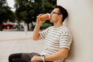 Young adult man sitting outdoors, drinking coffee, enjoying good weather and city view. Man has a break from work, spending time outside and relaxing. Time with yourself, dreaming, mental health. photo