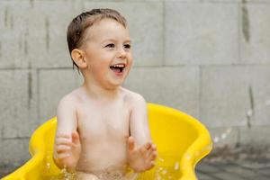 lindo niño bañándose en una bañera amarilla al aire libre. el niño feliz está salpicando, jugando con agua y divirtiéndose. temporada de verano y recreación. mantenerse fresco en el calor del verano. diversión acuática en el patio trasero. foto