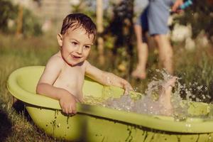 Cute little boy bathing in tub outdoors in garden. Happy child is splashing, playing with water and having fun. Summer season and recreation. Staying cool in the summer heat. Water fun in backyard. photo