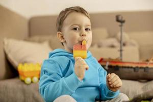 Little caucasian boy with blue hoodie playing with colourful toys and eating ice cream at home. Child having fun. Happy and cheerful kid plays with ship, dinosaurs, castle. Leisure activity. photo