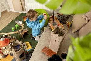 niño pequeño caucásico con capucha azul jugando con juguetes coloridos en casa. niño divirtiéndose. niño feliz y alegre juega con barco, dinosaurios, castillo. actividad de ocio, vida doméstica. habitación cómoda. foto