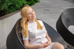Young beautiful blond woman sitting outdoors, on the wooden terrace in the city and resting. Girl has break, spending time outside and relaxing. Time with yourself, dreaming, relaxation, mental health photo