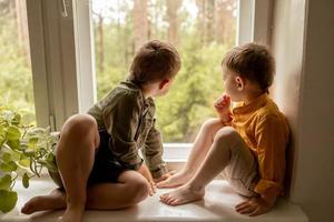 niños sentados en el alféizar de la ventana y esperando que alguien venga. dos hermanos, amigos. lindos niños en edad preescolar solos en casa. los niños están esperando a su madre o padre. soledad. padres ocupados. foto