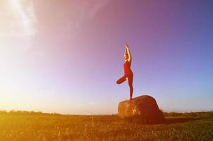 la silueta de una joven rubia con traje deportivo practica yoga en una pintoresca colina verde por la noche al atardecer. el concepto de ejercicio y estilos de vida saludables foto