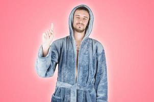 Portrait of young caucasian bearded man in blue bathrobe shows attention gesture isolated on pink background photo