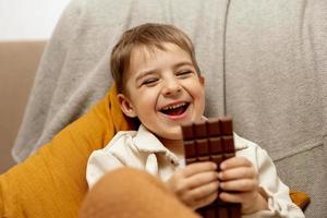 niño adorable sentado en el sofá de casa y comiendo barra de chocolate. niño y dulces, confitería de azúcar. niño disfrute de un delicioso postre. niño en edad preescolar con ropa informal. emoción positiva. foto