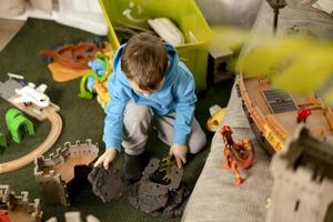 Little caucasian boy with blue hoodie playing with colourful toys at home. Child having fun. Happy and cheerful kid plays with ship, dinosaurs, castle. Leisure activity, domestic life. Cozy room. photo