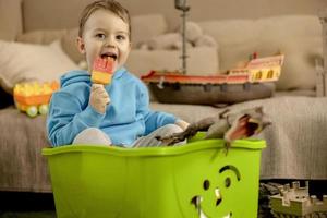 niño caucásico con capucha azul jugando con juguetes coloridos y comiendo helado en casa. niño divirtiéndose. niño feliz y alegre juega con barco, dinosaurios, castillo. actividad de ocio. foto