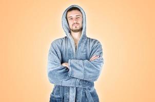 Portrait of young caucasian bearded man in blue bathrobe with crossed hands isolated on orange background photo