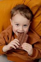 niño adorable sentado en el sofá de casa y comiendo barra de chocolate. niño y dulces, confitería de azúcar. niño disfrute de un delicioso postre. niño en edad preescolar con ropa informal. emoción positiva. foto