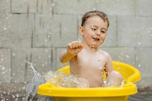 lindo niño bañándose en una bañera amarilla al aire libre. el niño feliz está salpicando, jugando con agua y divirtiéndose. temporada de verano y recreación. mantenerse fresco en el calor del verano. diversión acuática en el patio trasero. foto