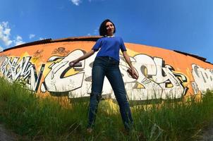 Portrait of an emotional young girl with black hair and piercings. A wide-angle photo of a girl with aerosol paint cans in the hands on a graffiti wall background. A modern portrait of a fisheye lens