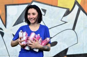 Portrait of an emotional young girl with black hair and piercings. Photo of a girl with aerosol paint cans in hands on a graffiti wall background. The concept of street art and use of aerosol paints