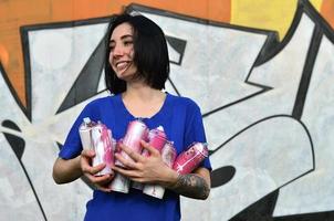 Portrait of an emotional young girl with black hair and piercings. Photo of a girl with aerosol paint cans in hands on a graffiti wall background. The concept of street art and use of aerosol paints