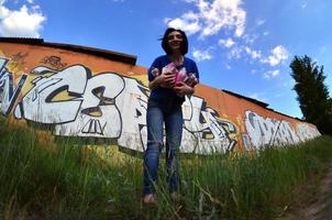 Portrait of an emotional young girl with black hair and piercings. A wide-angle photo of a girl with aerosol paint cans in the hands on a graffiti wall background. A modern portrait of a fisheye lens