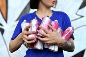 Portrait of an emotional young girl with black hair and piercings. Photo of a girl with aerosol paint cans in hands on a graffiti wall background. The concept of street art and use of aerosol paints