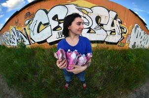 Portrait of an emotional young girl with black hair and piercings. A wide-angle photo of a girl with aerosol paint cans in the hands on a graffiti wall background. A modern portrait of a fisheye lens