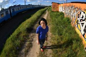 Portrait of an emotional young girl with black hair and piercings. A wide-angle photo of a girl with aerosol paint cans in the hands on a graffiti wall background. A modern portrait of a fisheye lens