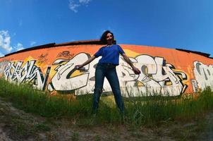 Portrait of an emotional young girl with black hair and piercings. A wide-angle photo of a girl with aerosol paint cans in the hands on a graffiti wall background. A modern portrait of a fisheye lens