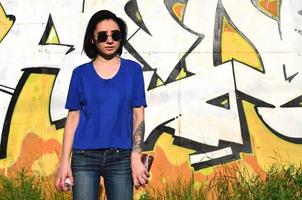 Portrait of an emotional young girl with black hair and piercings. Photo of a girl with aerosol paint cans in hands on a graffiti wall background. The concept of street art and use of aerosol paints