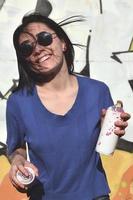Portrait of an emotional young girl with black hair and piercings. Photo of a girl with aerosol paint cans in hands on a graffiti wall background. The concept of street art and use of aerosol paints
