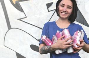 Portrait of an emotional young girl with black hair and piercings. Photo of a girl with aerosol paint cans in hands on a graffiti wall background. The concept of street art and use of aerosol paints