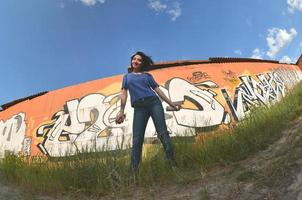 Portrait of an emotional young girl with black hair and piercings. A wide-angle photo of a girl with aerosol paint cans in the hands on a graffiti wall background. A modern portrait of a fisheye lens