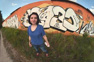 Portrait of an emotional young girl with black hair and piercings. A wide-angle photo of a girl with aerosol paint cans in the hands on a graffiti wall background. A modern portrait of a fisheye lens