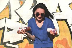 Portrait of an emotional young girl with black hair and piercings. Photo of a girl with aerosol paint cans in hands on a graffiti wall background. The concept of street art and use of aerosol paints